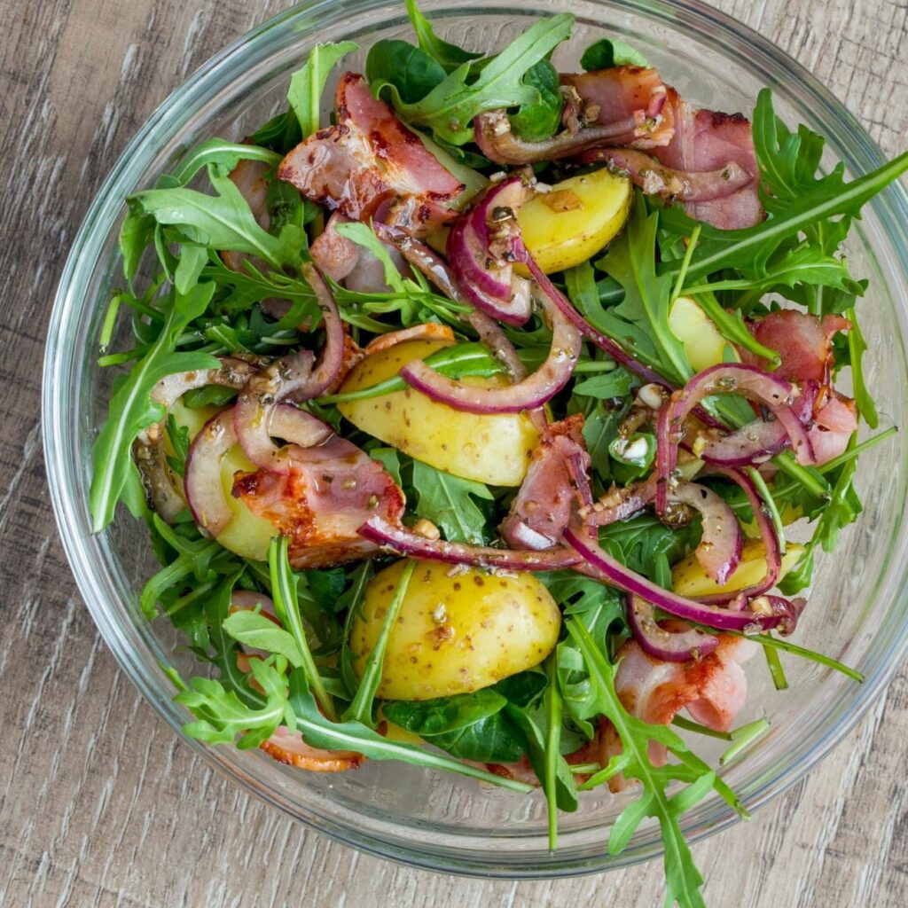 Kartoffel-Rucola-Salat wird von oben in einer Glasschüssel präsentiert.