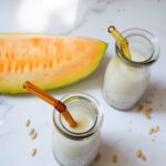 Melonenkernmilch wir in zwei kleinen Flaschen mit Strohhalmen von oben mit einem Stück Cantaloupe-Melone gezeigt.